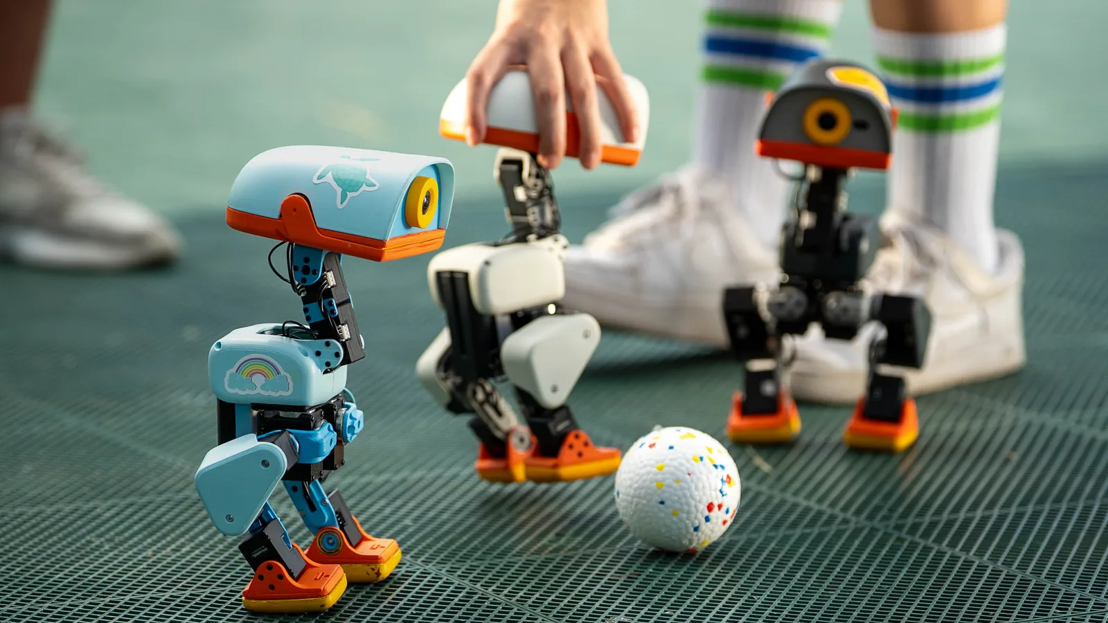 Three Microduck robots in sky blue, cream and graphite shells around a ball on a court floor, one lifted by a hand